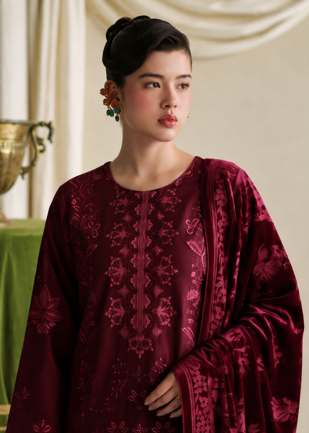 A woman wearing a deep burgundy embroidered traditional outfit with a matching draped shawl, set against a soft beige draped background with a gold urn prop.