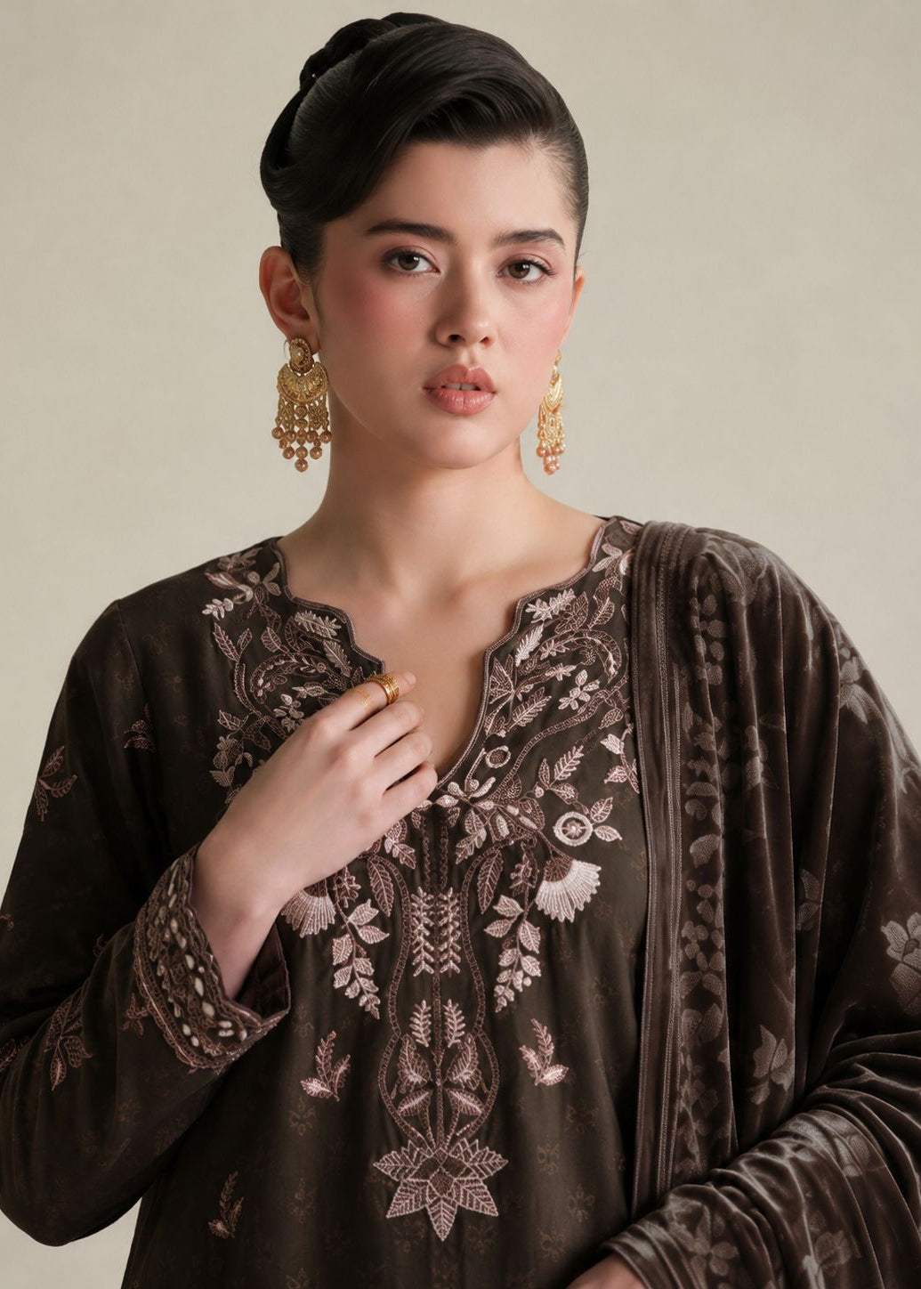 A woman wearing a dark brown embroidered kurta with floral and geometric patterns, paired with a matching brown dupatta and gold earrings, set against a plain beige background.