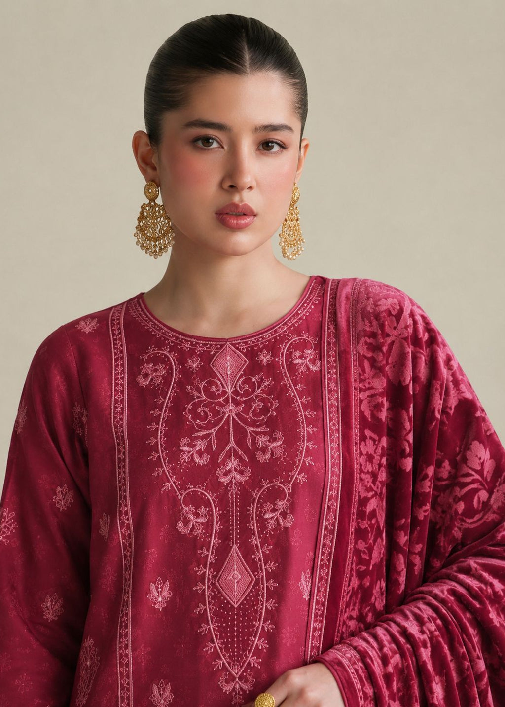 A woman wearing a deep magenta velvet embroidered kurta with a matching dupatta and ornate gold earrings, set against a solid beige background.