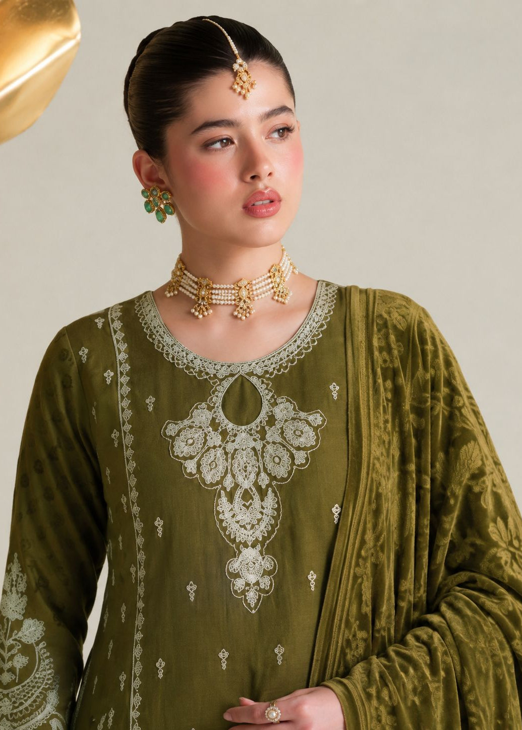 A woman wearing an olive green traditional embroidered outfit with white floral embroidery on the neckline and sleeves, styled with gold jewelry against a light beige background.