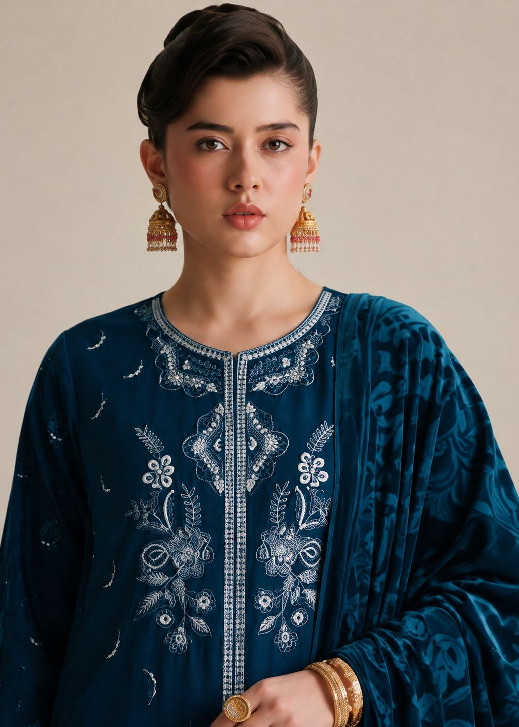 A woman wearing a dark teal blue embroidered kurta with white floral embroidery and a matching patterned dupatta, styled with gold earrings and bracelets against a neutral beige background.