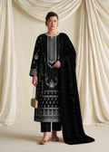 A woman stands in front of draped cream fabric, wearing a black traditional outfit with ornate silver embroidery and a long black embroidered dupatta. She holds a small beige handbag and wears beige flat shoes.