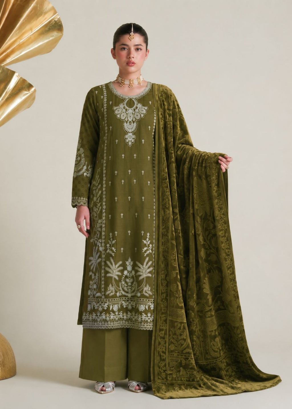 A woman models an olive green traditional South Asian outfit with white embroidery, matching wide-leg trousers, and a sheer embroidered dupatta, posed against a neutral beige background with decorative gold props.