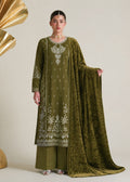 A woman models an olive green traditional South Asian outfit with white embroidery, matching wide-leg trousers, and a sheer embroidered dupatta, posed against a neutral beige background with decorative gold props.