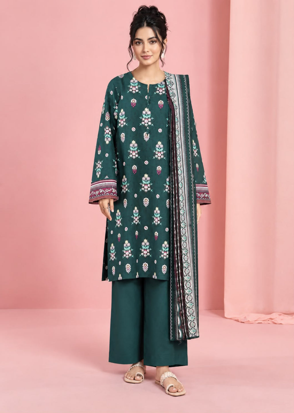 A woman models a three-piece dark teal green ethnic suit set featuring a printed kurta, matching wide-leg trousers, and a patterned dupatta, set against a pink studio backdrop.