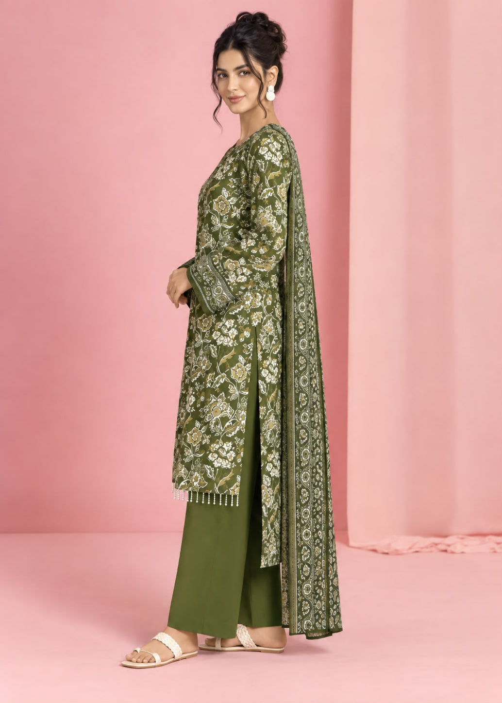 A woman models an olive green traditional outfit with intricate gold and white floral embroidery, paired with matching olive green pants, set against a solid pink studio backdrop.