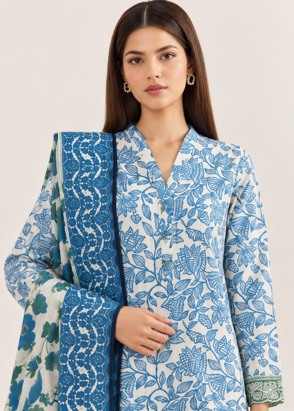 A woman modeling a white three-piece ethnic suit set featuring a blue floral print kurta, matching trousers, and a coordinating dupatta with intricate blue and green border patterns, set against a plain beige background.