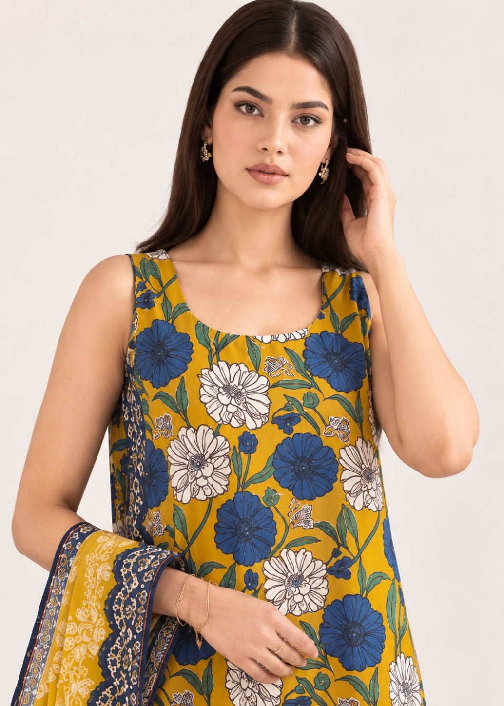 A woman modeling a sleeveless mustard yellow kurta with a blue and white floral print, paired with a matching patterned dupatta, against a light gray studio background.