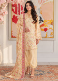 A woman models a traditional three-piece salwar kameez suit in a warm beige/cream color with a delicate pink and yellow floral print, paired with a matching printed dupatta featuring a pink border, set against an indoor background with a framed abstract painting.