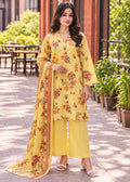 A woman modeling a yellow floral salwar kameez suit with matching wide-leg pants and a sheer printed dupatta, standing outdoors on a paved walkway with plants and tables in the background.