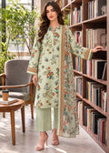 A woman models a light sage green three-piece salwar kameez suit with a floral print and orange border detailing, standing in a cozy room with a bookshelf background.
