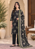 A woman models a traditional three-piece salwar kameez suit set in a courtyard setting. The set features a black kurta with a beige and green floral print, black wide-leg trousers, and a matching dupatta with a green border and beige floral print.
