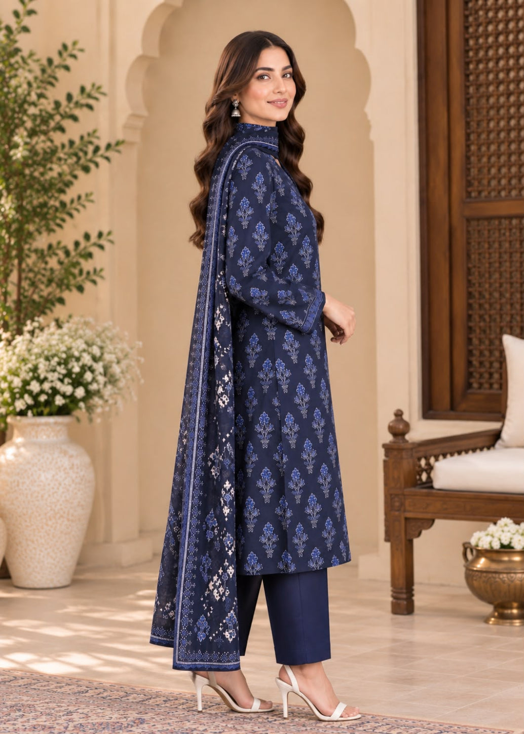 A woman models a navy blue three-piece traditional outfit with matching dupatta, featuring intricate blue and white floral prints, set against an elegant indoor architectural background.