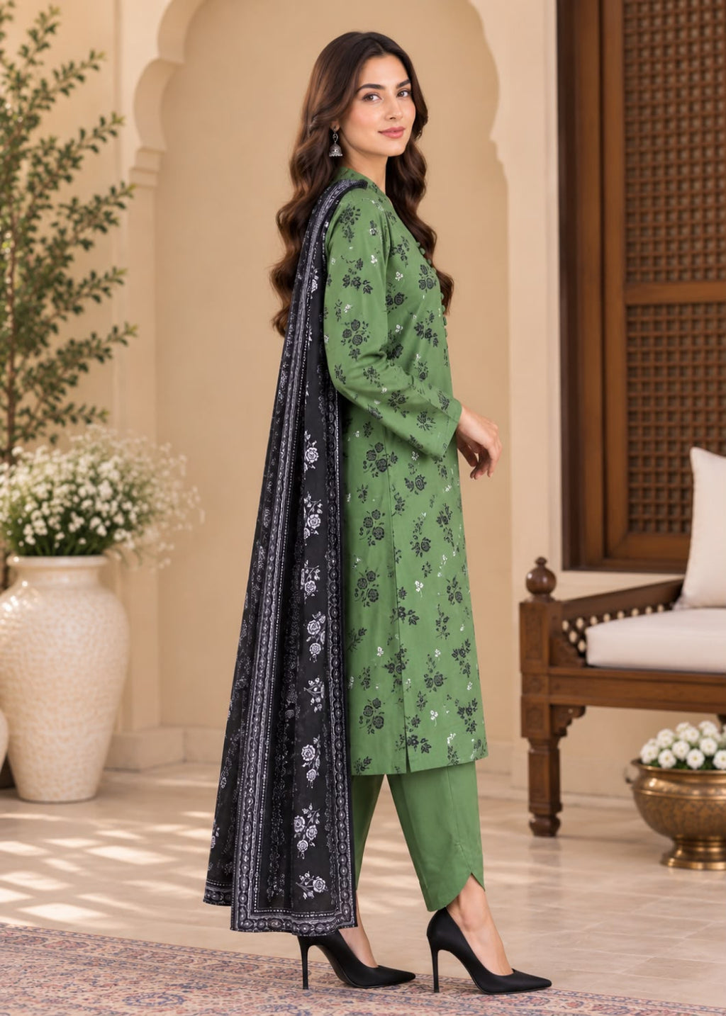 A woman models a green three-piece ethnic suit with black floral embroidery, matching green pants, and a black dupatta with white border and floral prints, set against a warm, traditional architectural background.