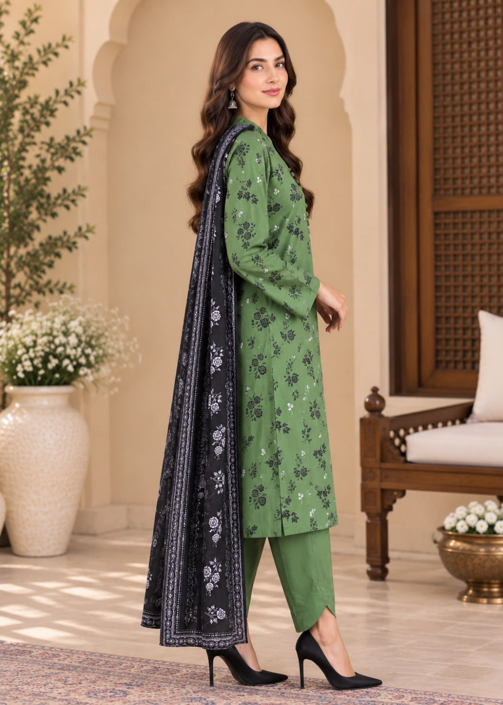 A woman models a green three-piece ethnic suit with black floral embroidery, matching green pants, and a black dupatta with white border and floral prints, set against a warm, traditional architectural background.