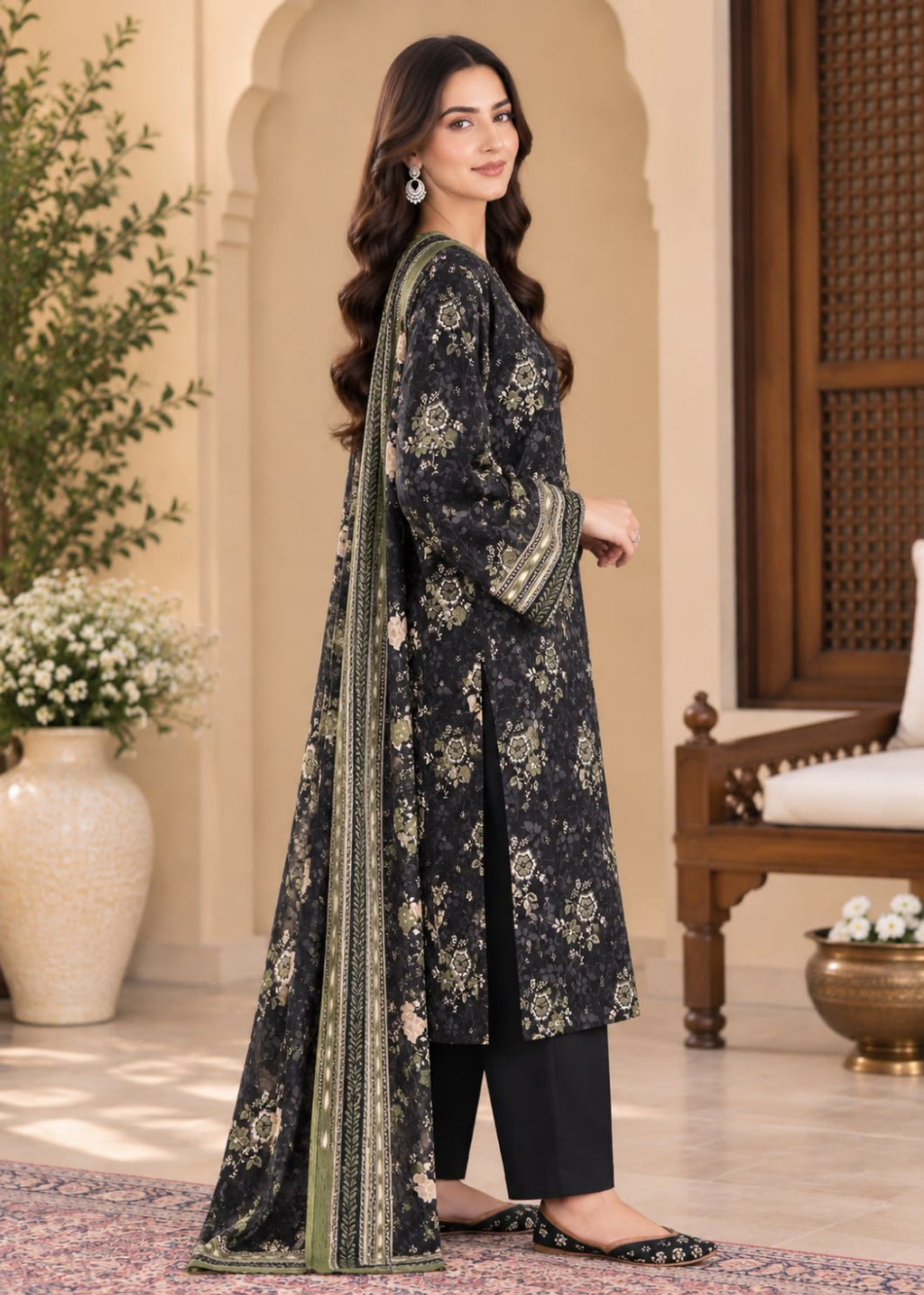 A woman models a black three-piece traditional outfit with beige floral prints and green border details, consisting of a long kurta, pants, and a dupatta, set against an elegant indoor architectural background.