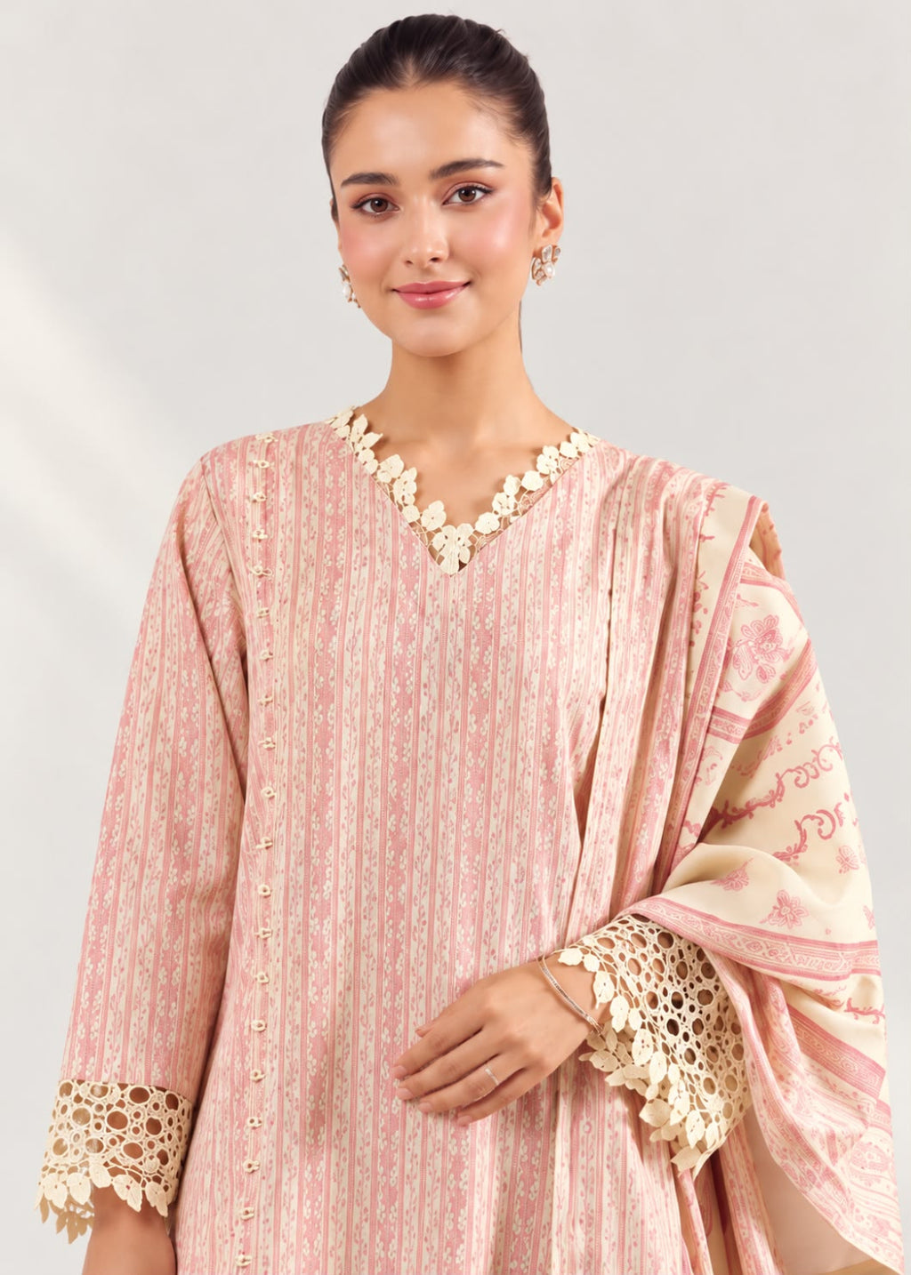 A woman modeling a light pink traditional outfit with vertical stripes, cream lace trim at the neckline and cuffs, and a matching printed dupatta draped over one shoulder, against a plain light gray studio background.