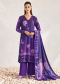 A woman models a purple three-piece ethnic suit with floral prints and a matching dupatta, standing on a bed of white pebbles against a light studio backdrop.