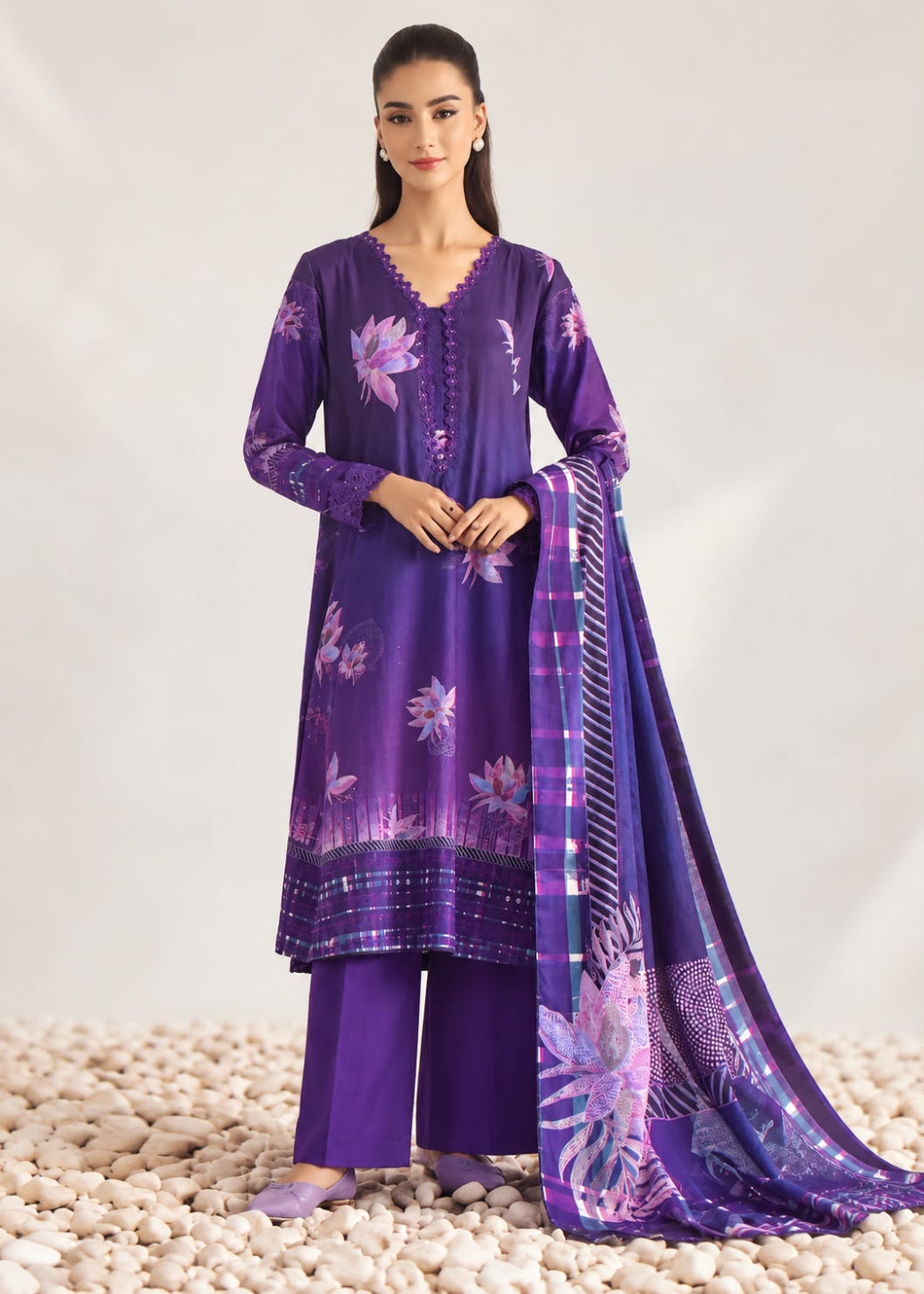 A woman models a purple three-piece ethnic suit with floral prints and a matching dupatta, standing on a bed of white pebbles against a light studio backdrop.