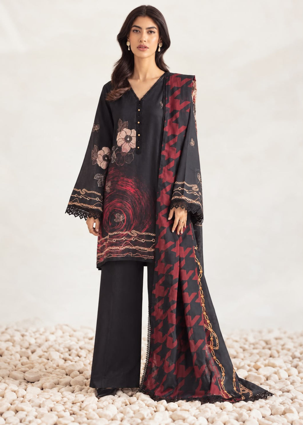 A woman models a black three-piece traditional outfit with red and gold floral patterns, featuring a long kurta, wide-leg trousers, and a matching dupatta, set against a neutral studio backdrop with decorative pebbles.
