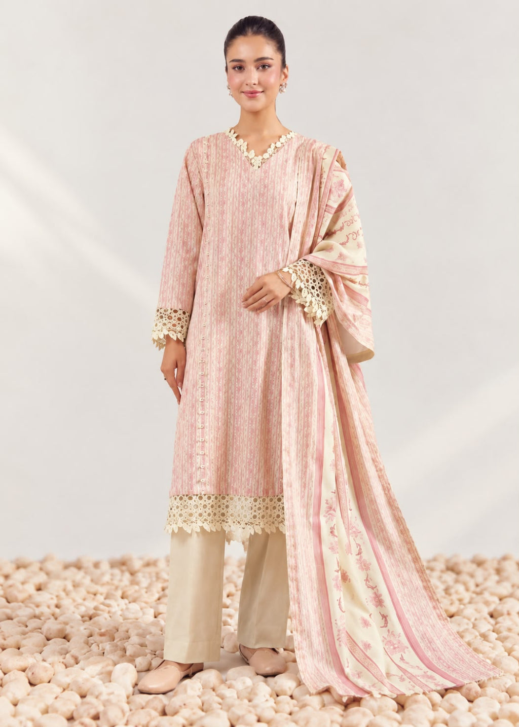 A woman models a traditional three-piece salwar kameez suit in light pink and beige. The kurta features vertical stripes, a V-neckline with a gold embroidered border, and lace-trimmed cuffs.