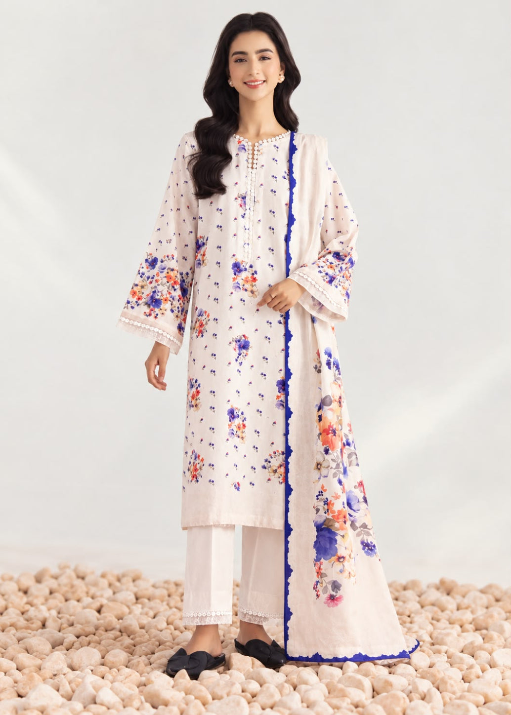 A woman models a traditional three-piece salwar kameez suit in off-white, featuring colorful floral embroidery in blue, orange, and purple, with a matching dupatta and white trousers, standing on a bed of light-colored pebbles.