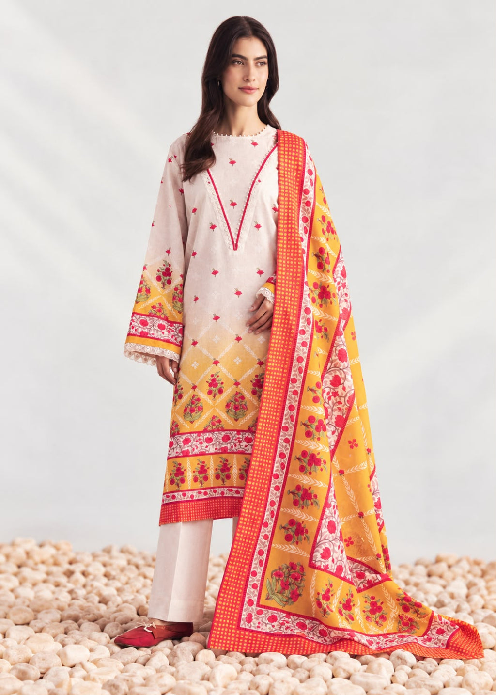 A woman models a traditional three-piece salwar kameez suit in cream and yellow, featuring red floral embroidery, a red-orange border, and a matching printed dupatta, set against a light gray studio backdrop with decorative pebbles at the bottom.
