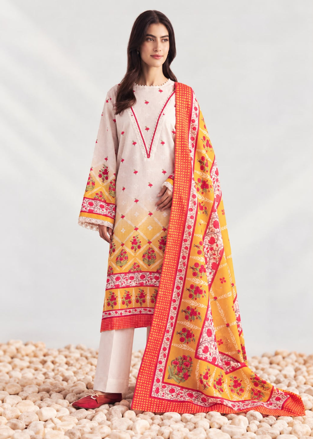 A woman models a traditional three-piece salwar kameez suit in cream and yellow, featuring red floral embroidery, a red-orange border, and a matching printed dupatta, set against a light gray studio backdrop with decorative pebbles at the bottom.