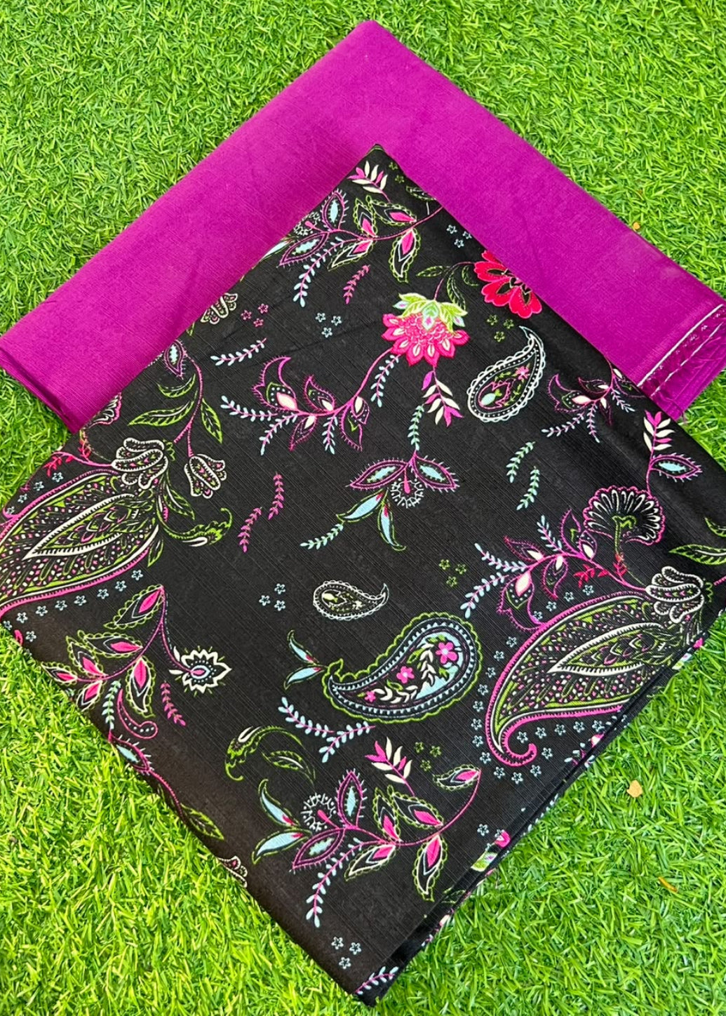 A folded black fabric piece with a colorful paisley and floral print, placed on bright green artificial grass with a folded magenta fabric piece behind it.