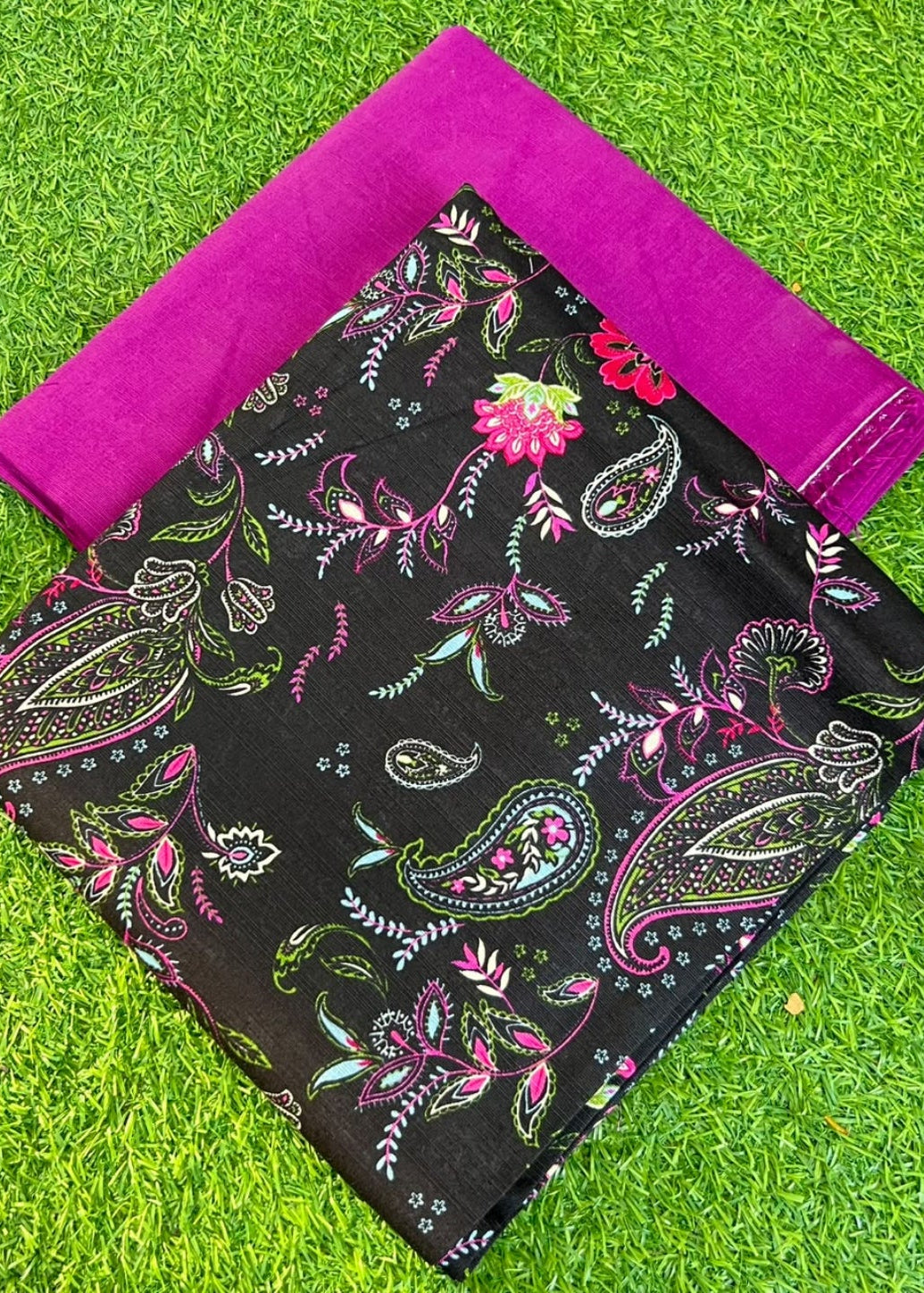 A folded black fabric piece with a colorful paisley and floral print, placed on bright green artificial grass with a folded magenta fabric piece behind it.