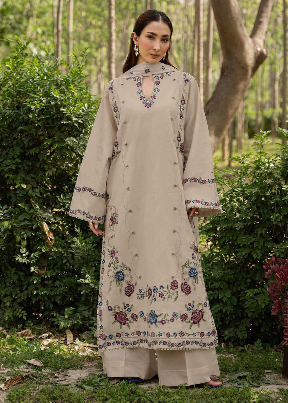 A woman stands outdoors in a wooded garden setting wearing a long beige embroidered traditional outfit with wide sleeves and floral detailing in blue, pink, and green.