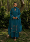 A woman stands outdoors in a wooded setting wearing a teal traditional outfit with floral embroidery and a matching dupatta.
