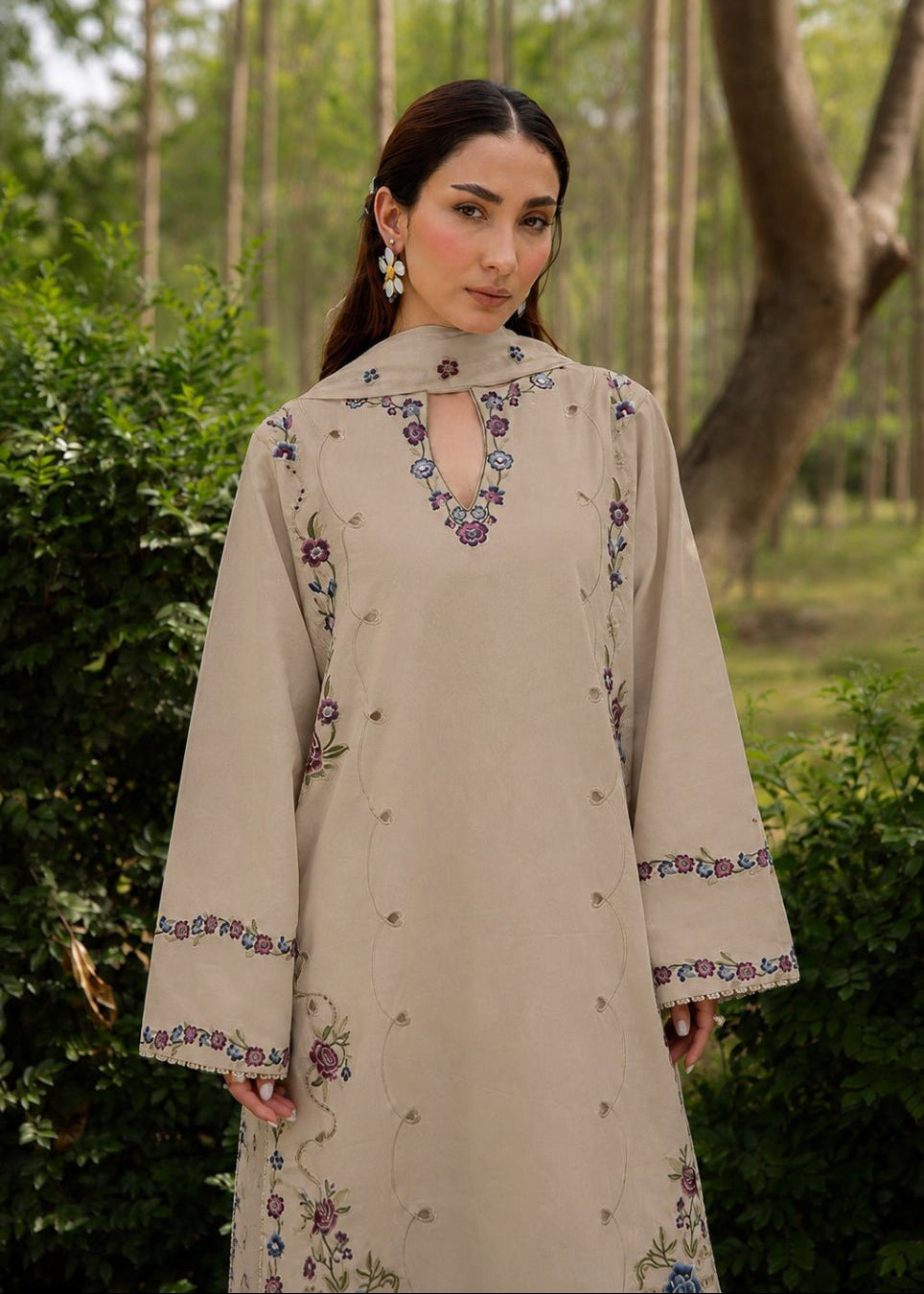 A woman wearing a beige long-sleeved embroidered dress or coat with floral detailing, standing outdoors in front of a blurred background of trees and greenery.