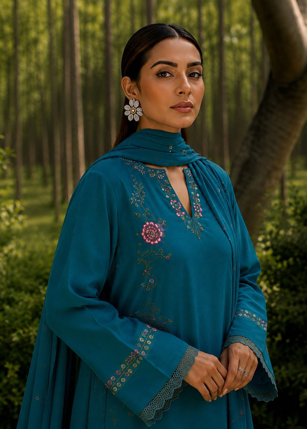 A woman wearing a teal blue embroidered kurta with a matching dupatta and white floral earrings, standing in a wooded outdoor setting.