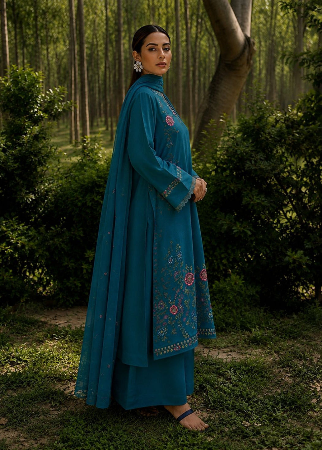 A woman wearing a teal blue embroidered traditional outfit with a matching dupatta, standing in a dense forest setting.