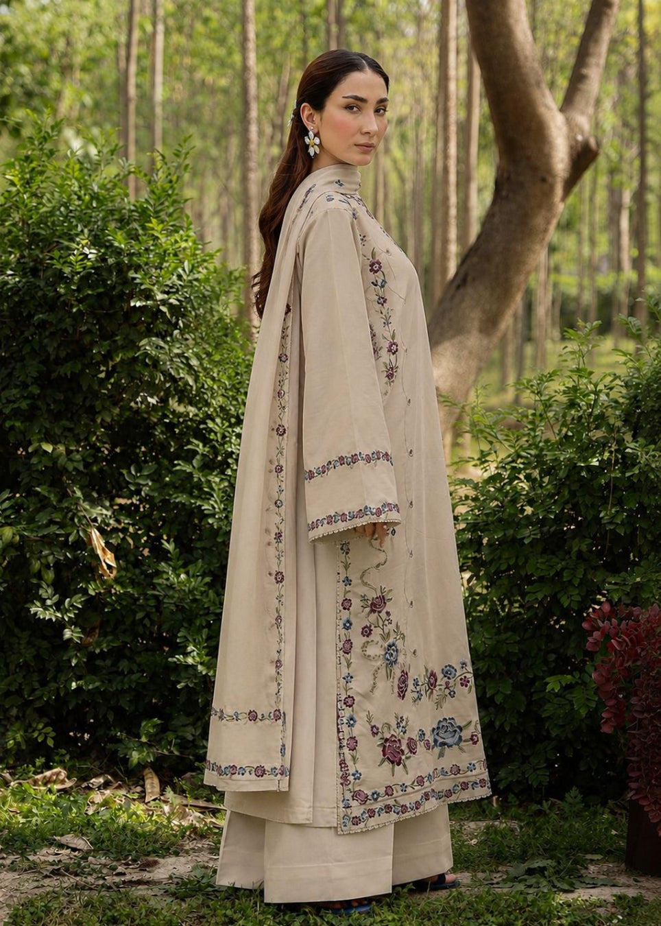 A woman stands outdoors in a wooded garden setting wearing a long beige embroidered traditional outfit with wide sleeves and matching pants. The garment has floral and geometric border embroidery in muted blue, purple, and green tones.