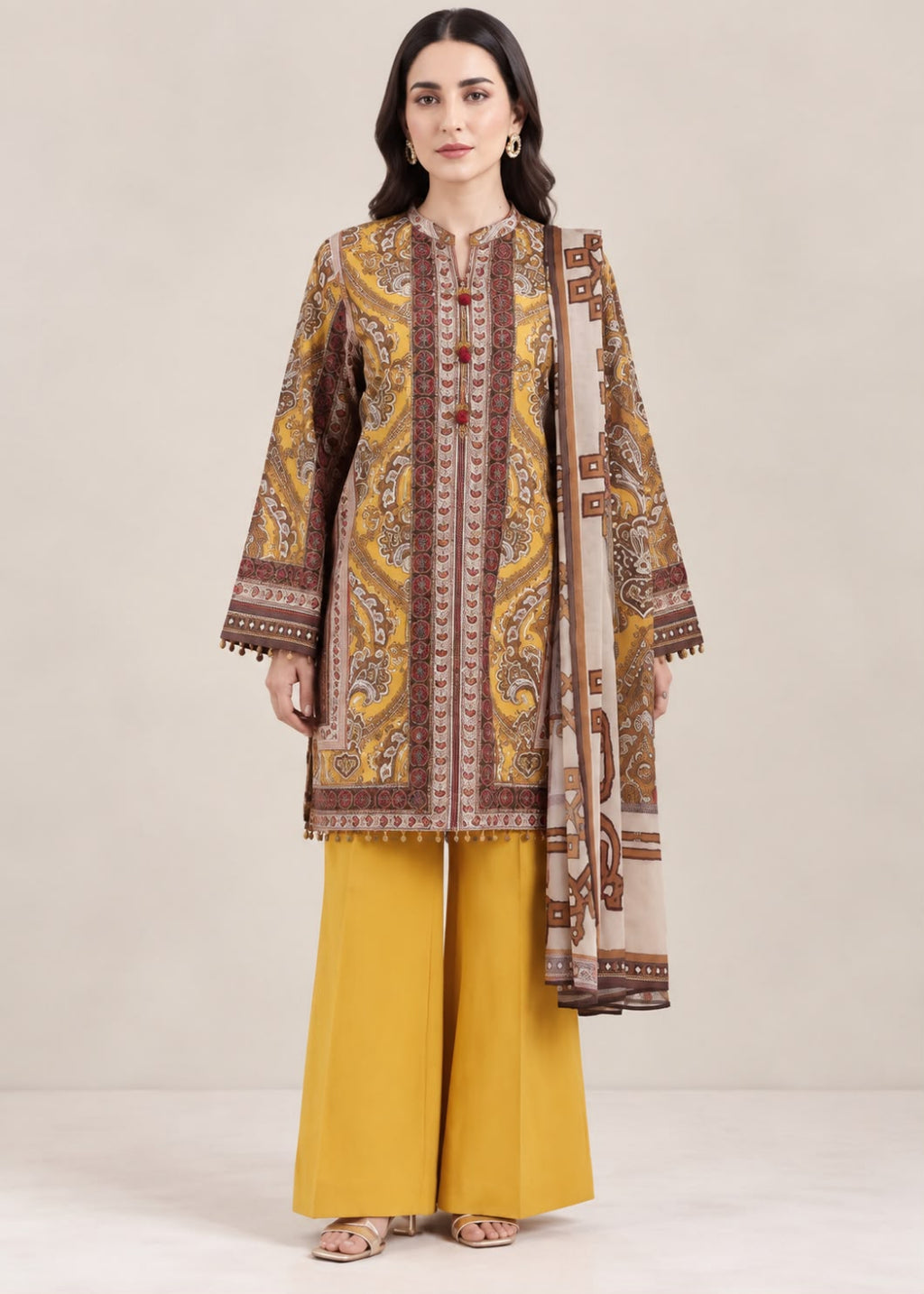 A woman modeling a traditional three-piece outfit featuring a mustard yellow kurta with brown and beige paisley patterns, matching wide-leg trousers, and a coordinating dupatta, set against a plain beige studio background.