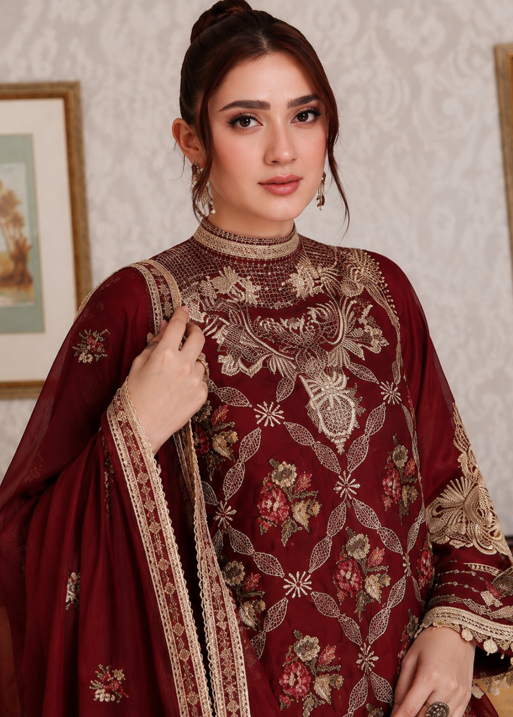 A woman wearing a traditional maroon and gold embroidered ethnic outfit with a matching dupatta, posed in front of a patterned wallpaper background with framed art.