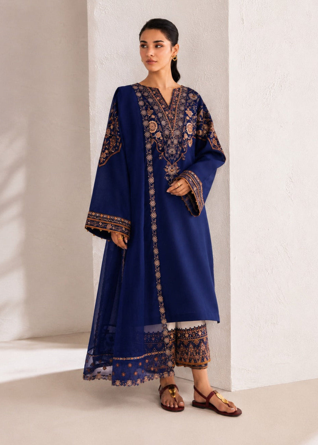 A woman models a navy blue traditional outfit with intricate gold and brown embroidery, consisting of a long kurta, matching dupatta, and patterned trousers, set against a minimalist beige wall and floor.