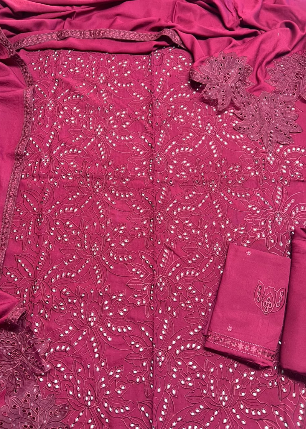 A close-up of a magenta pink embroidered fabric, likely a traditional salwar suit material, featuring intricate floral embroidery, beadwork, and a matching fabric pocket with embroidered detailing.