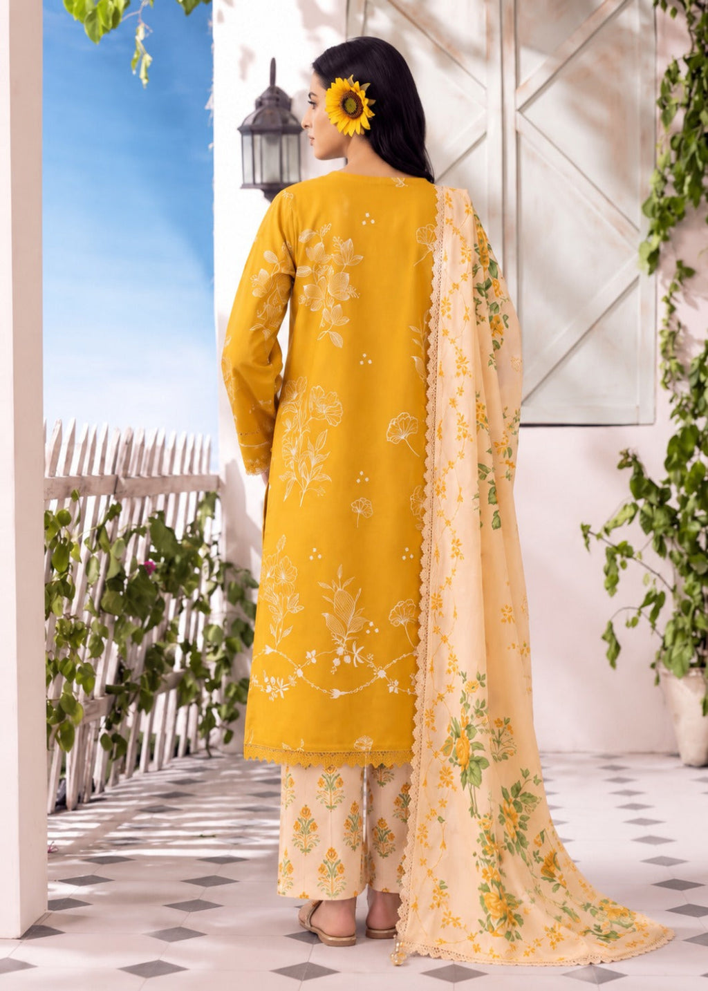 A woman stands with her back to the camera wearing a mustard yellow traditional outfit with white floral embroidery, beige patterned pants, and a matching beige dupatta with green and yellow floral print. She is posed in a bright, sunlit outdoor-style setting with a white fence, blue sky backdrop, and greenery.