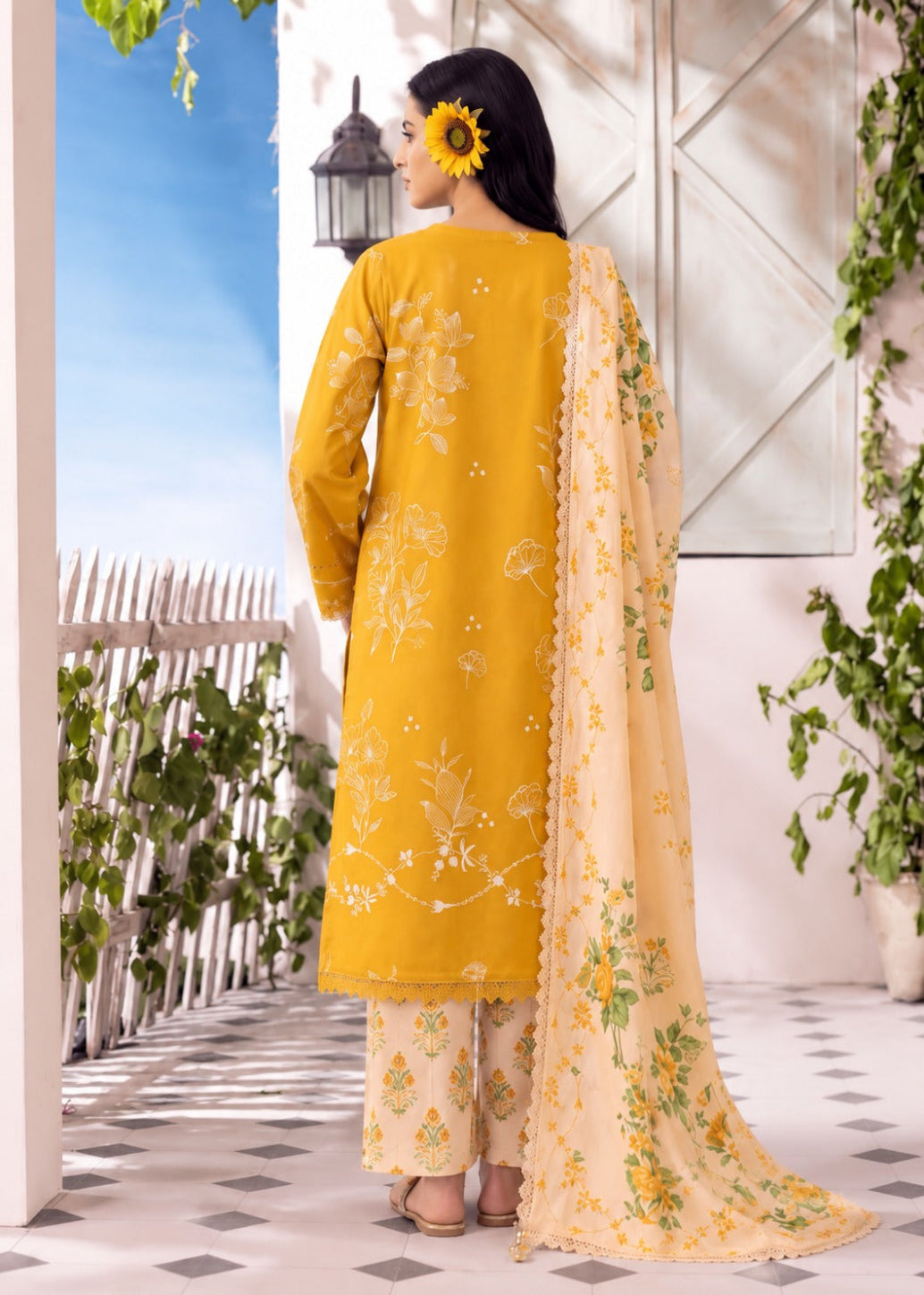 A woman stands with her back to the camera wearing a mustard yellow traditional outfit with white floral embroidery, beige patterned pants, and a matching beige dupatta with green and yellow floral print. She is posed in a bright, sunlit outdoor-style setting with a white fence, blue sky backdrop, and greenery.