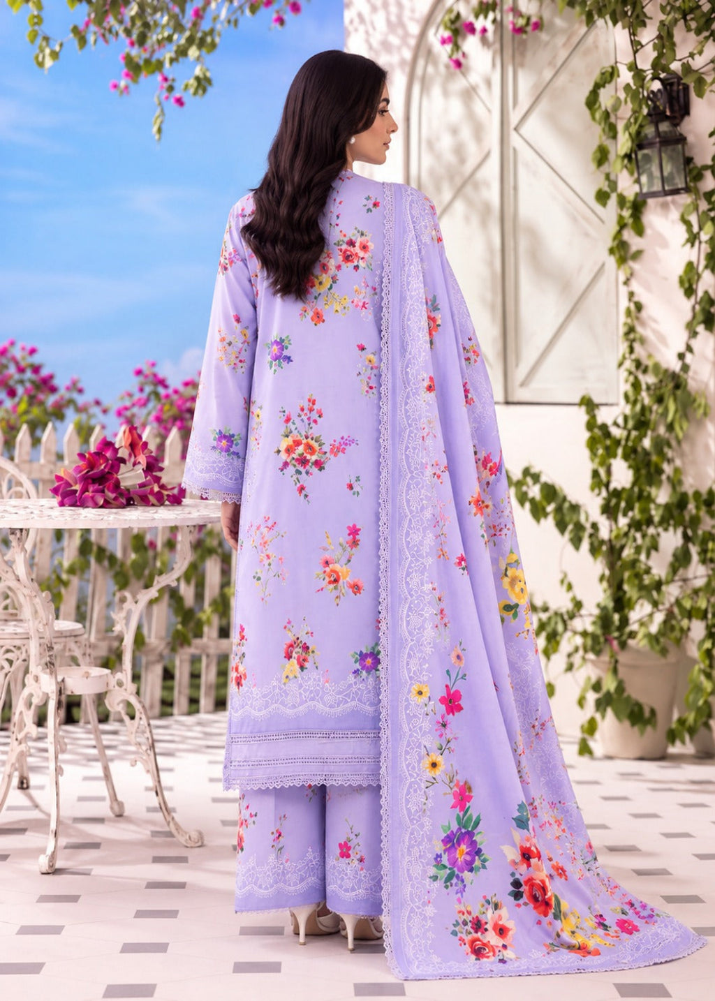 A woman stands outdoors wearing a lavender three-piece traditional outfit with colorful floral embroidery and a matching dupatta. The scene includes a patio, white table and chairs, blue sky, and greenery.
