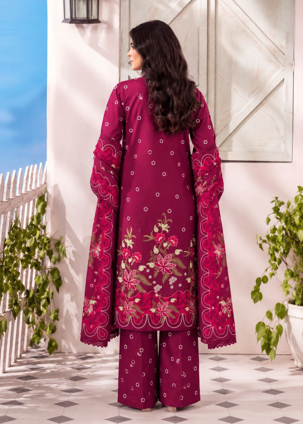 A woman stands outdoors wearing a deep magenta three-piece salwar kameez suit with intricate floral embroidery and mirror work on the sleeves and back. The outfit includes a long tunic, wide collar, and matching trousers, set against a light-colored stucco wall with potted plants.