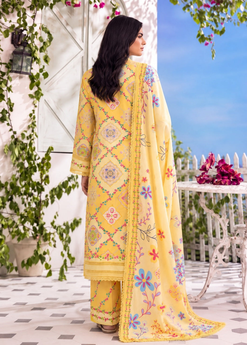 A woman stands on a balcony wearing a bright yellow traditional outfit with colorful floral embroidery and a matching dupatta. The setting features a white wall with green vines, a blue sky, and a white table with flowers.