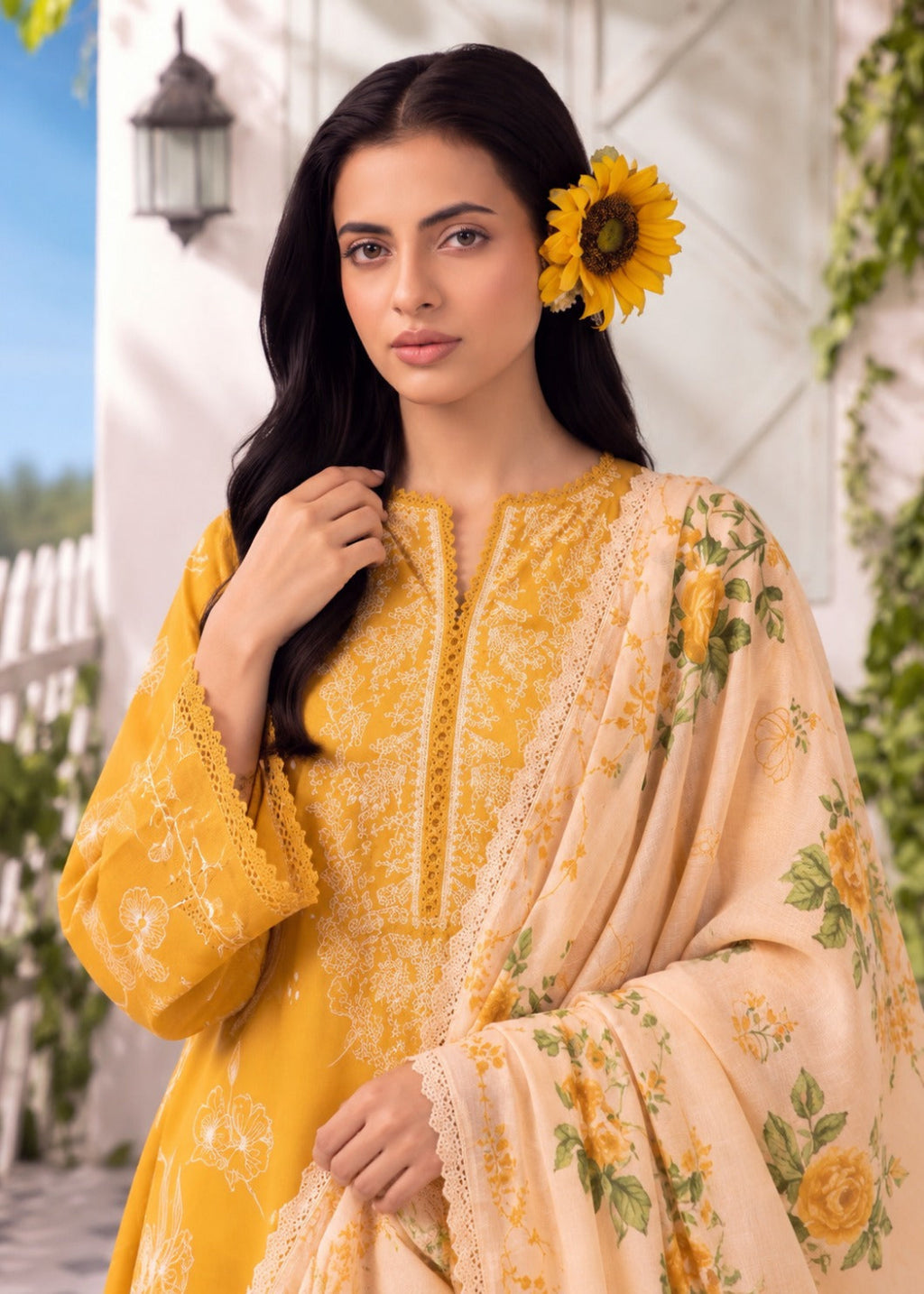 A woman modeling a traditional three-piece salwar kameez suit in mustard yellow and beige. The kurta features intricate yellow embroidery, paired with a beige dupatta decorated with yellow floral prints and green leaves.