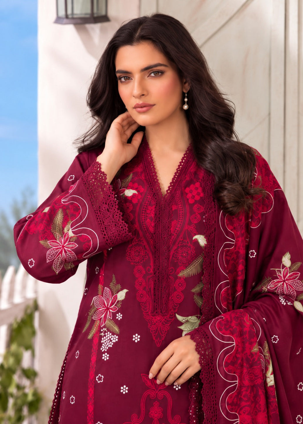 A woman wearing a deep maroon embroidered traditional outfit with floral patterns and lace sleeves, posed against a light-colored outdoor wall.