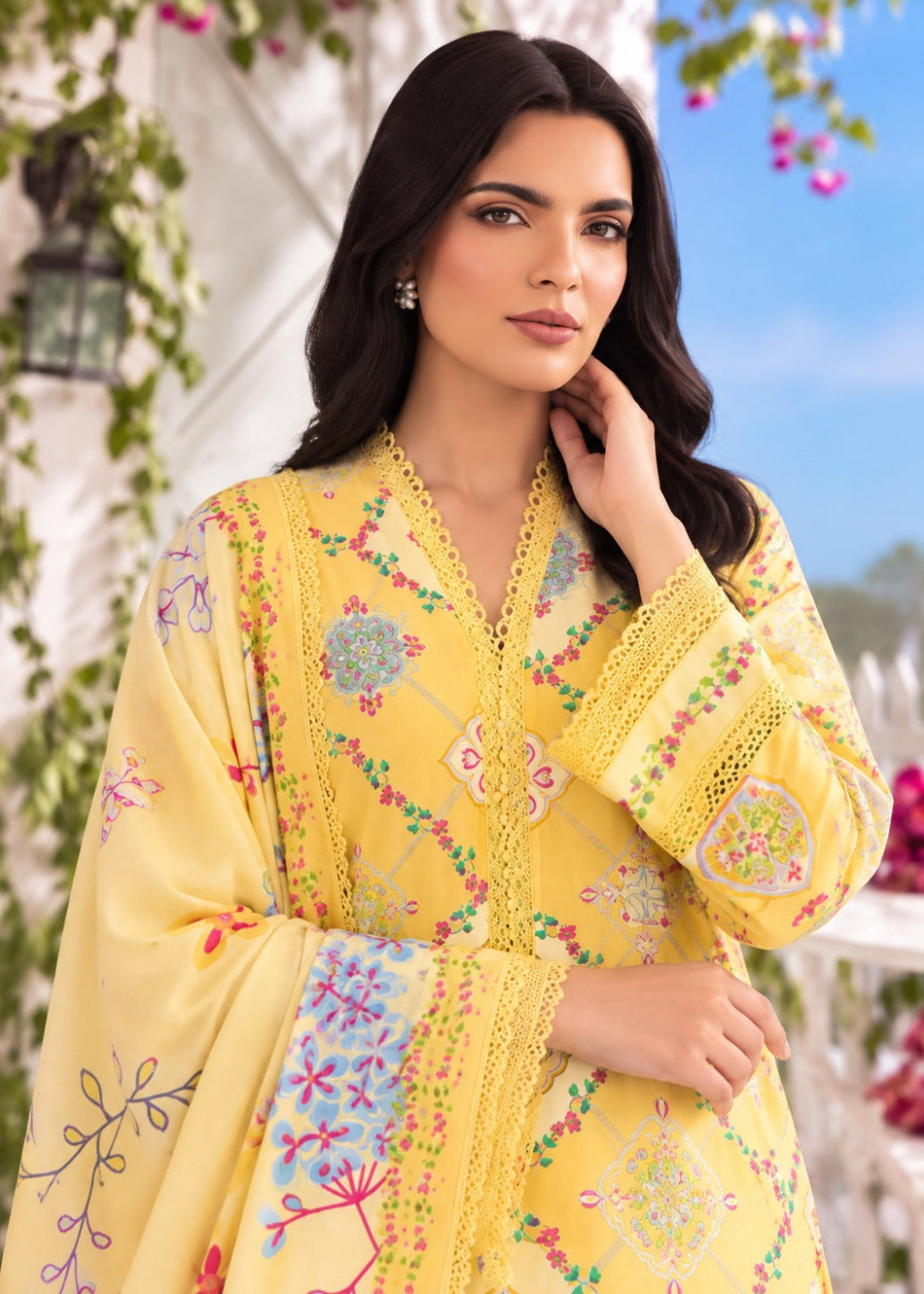 A woman wearing a bright yellow embroidered traditional outfit with floral patterns and lace trim, posed outdoors against a soft-focus background of greenery and blue sky.
