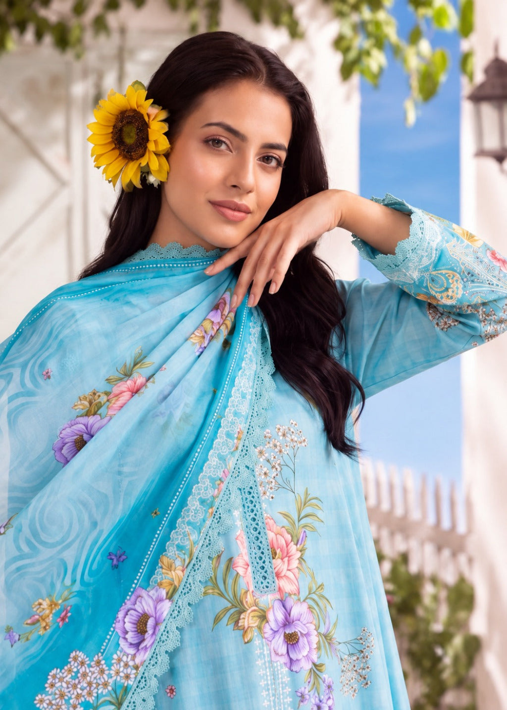 A woman wearing a light blue embroidered traditional outfit with a matching dupatta, featuring floral prints in pink, purple, and green. She has a yellow sunflower tucked into her dark hair, posing outdoors against a white architectural background with green foliage.