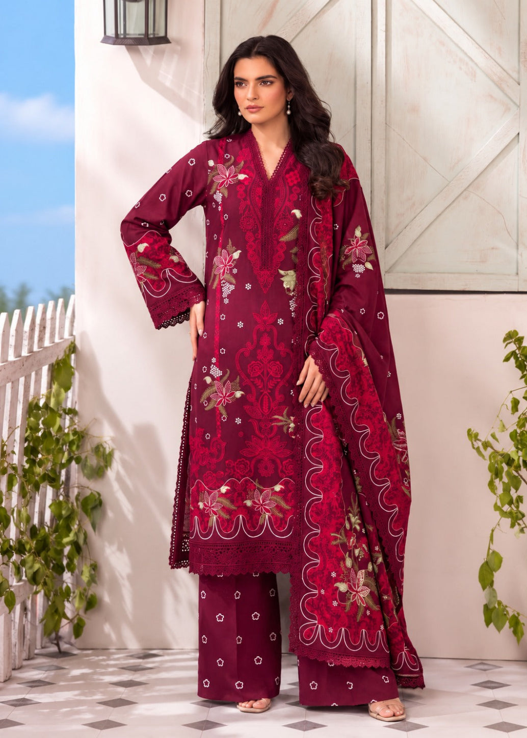 A woman models a deep magenta three-piece traditional outfit with intricate floral embroidery and gold detailing, set against a rustic white wall and tiled floor.